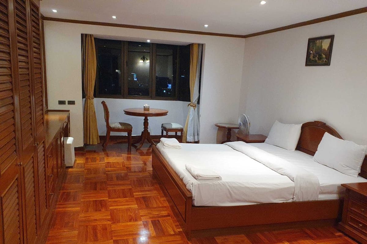 A hotel room with a wooden floor, a bed with white linens, a small round table with a chair, a window with curtains, and a nightstand with a fan.