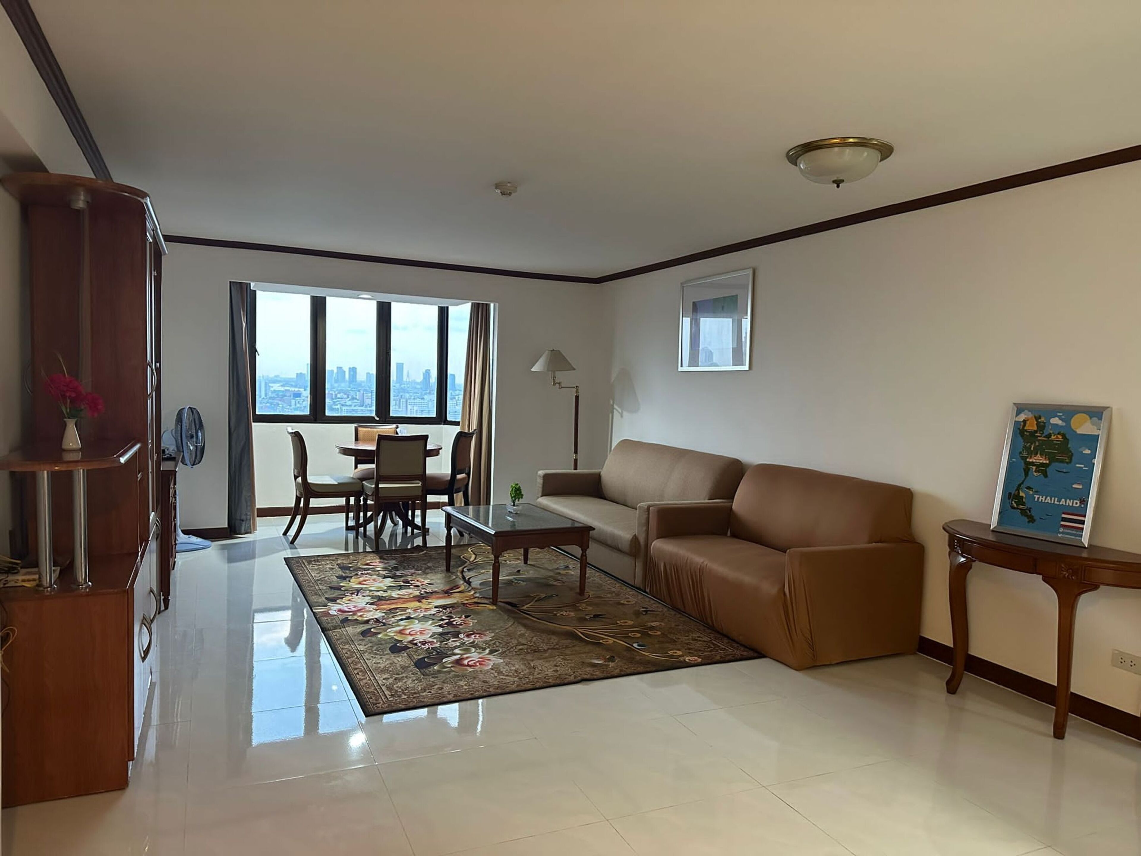 A modern living room with a sofa, a coffee table, and a dining area with chairs. There is a large window providing a view of the city.
