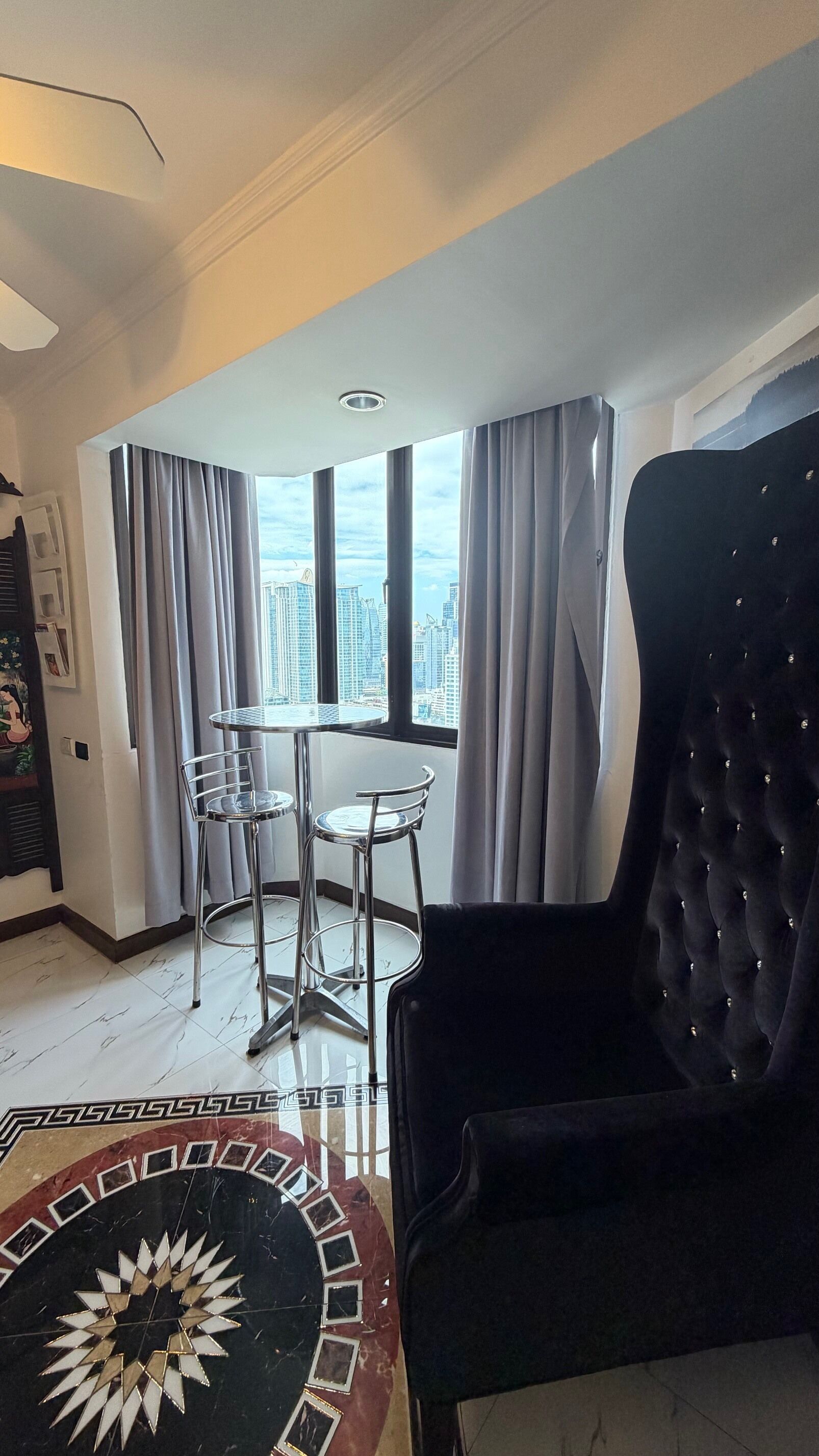 Un salon moderne doté d’une grande fenêtre, d’une table ronde avec deux chaises, d’un fauteuil noir à capitonnage et d’un tapis à motifs.
