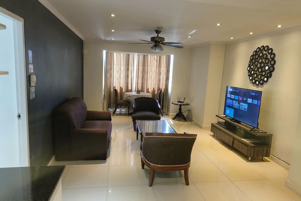 A modern living room with a sofa, armchair, and coffee table. A TV is mounted on the wall, and a ceiling fan is visible.