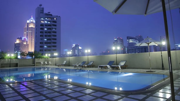 A rooftop pool with lounge chairs and umbrellas, surrounded by city buildings at dusk. - Omni Tower Sukhumvit Nana - Direct (Bangkok)