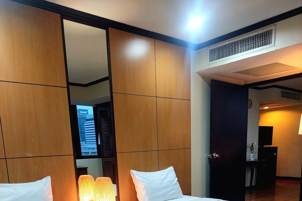 A hotel room with two beds, a wooden wardrobe, a desk with a lamp, and a view of the cityscape through the window.