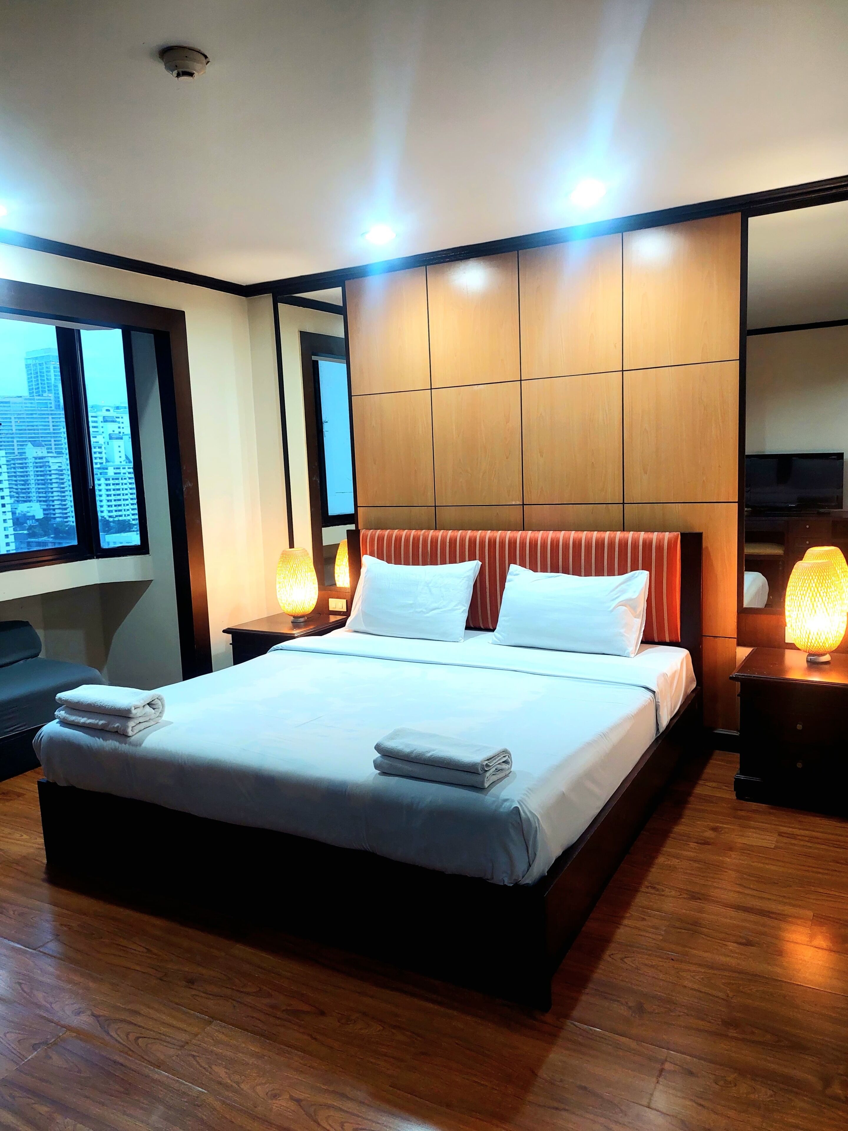 A hotel room with a large bed, wooden flooring, a city view through the window, and a modern design.