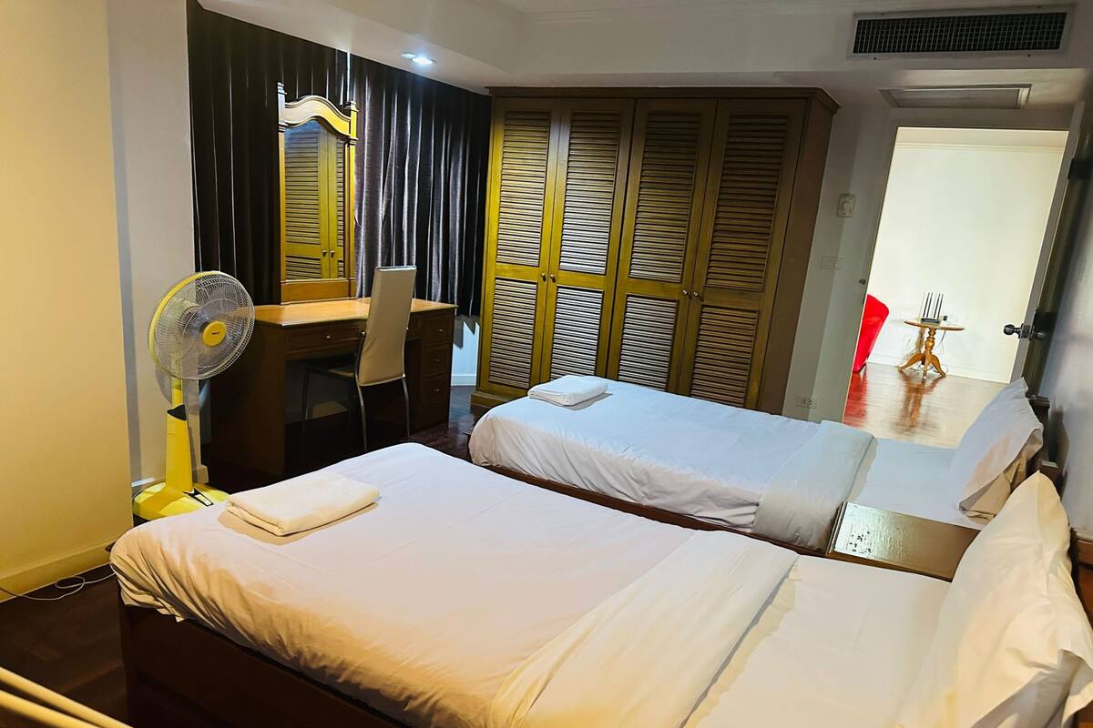 A hotel room with two beds, a desk, a chair, a fan, and a wardrobe with louvered doors.