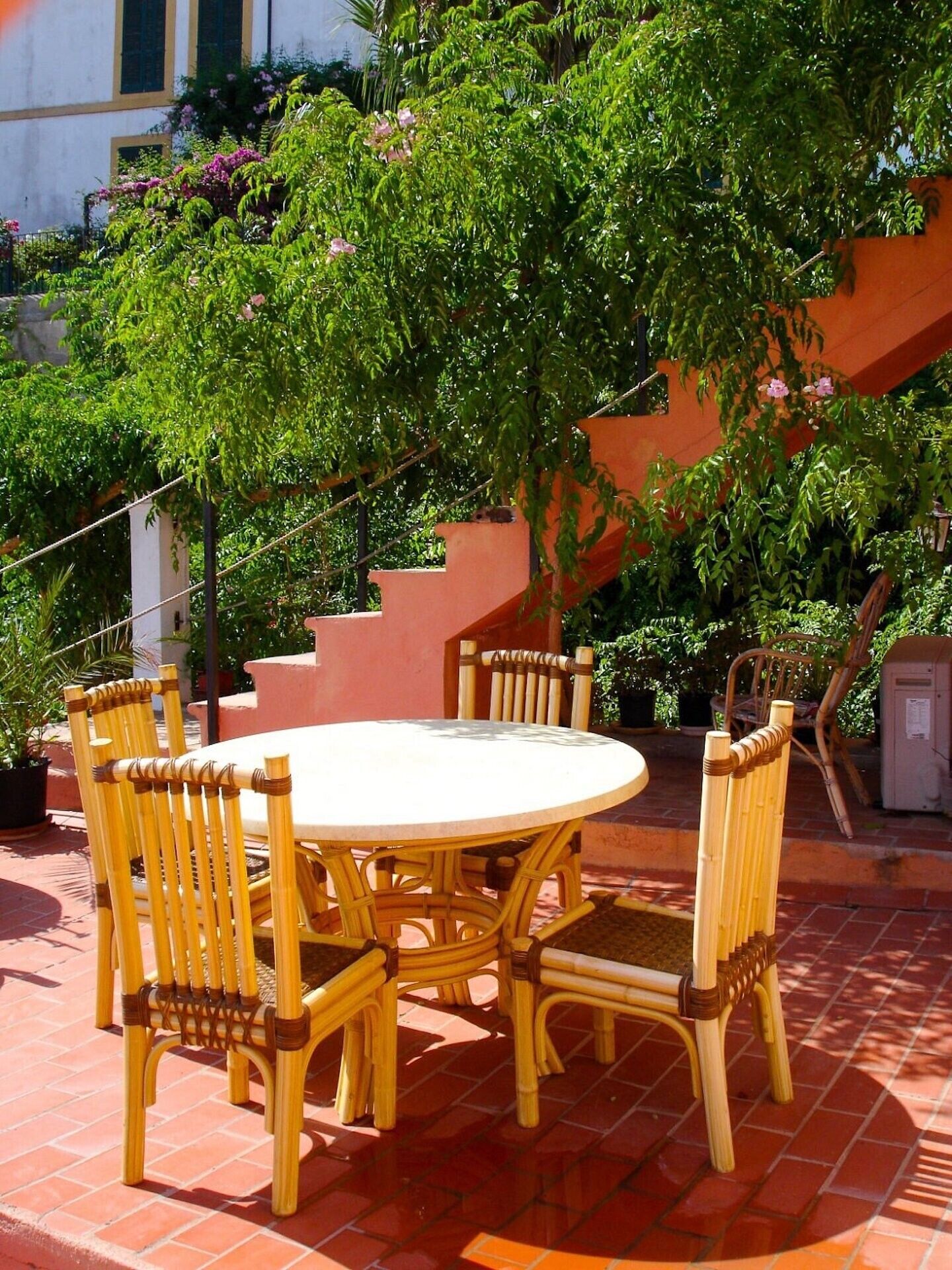 A round table with six chairs, surrounded by lush greenery and a staircase.