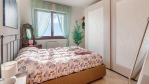 Ein Schlafzimmer mit Bett, Heizkörper, Fenster mit durchsichtigen Vorhängen, Spiegel und Kleiderschrank.