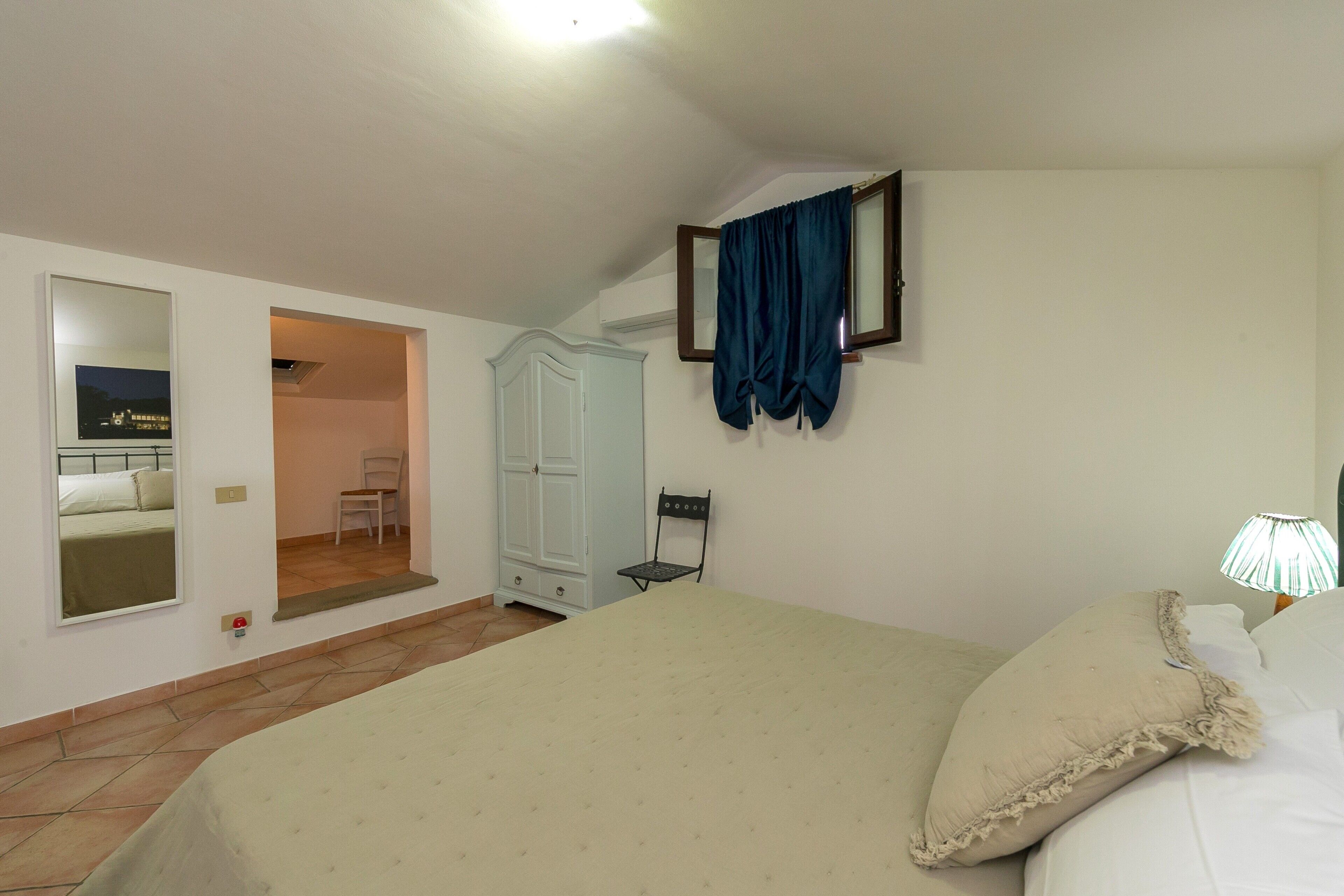 A bedroom with a bed, a wardrobe, a chair, a mirror, and a door leading to another room.