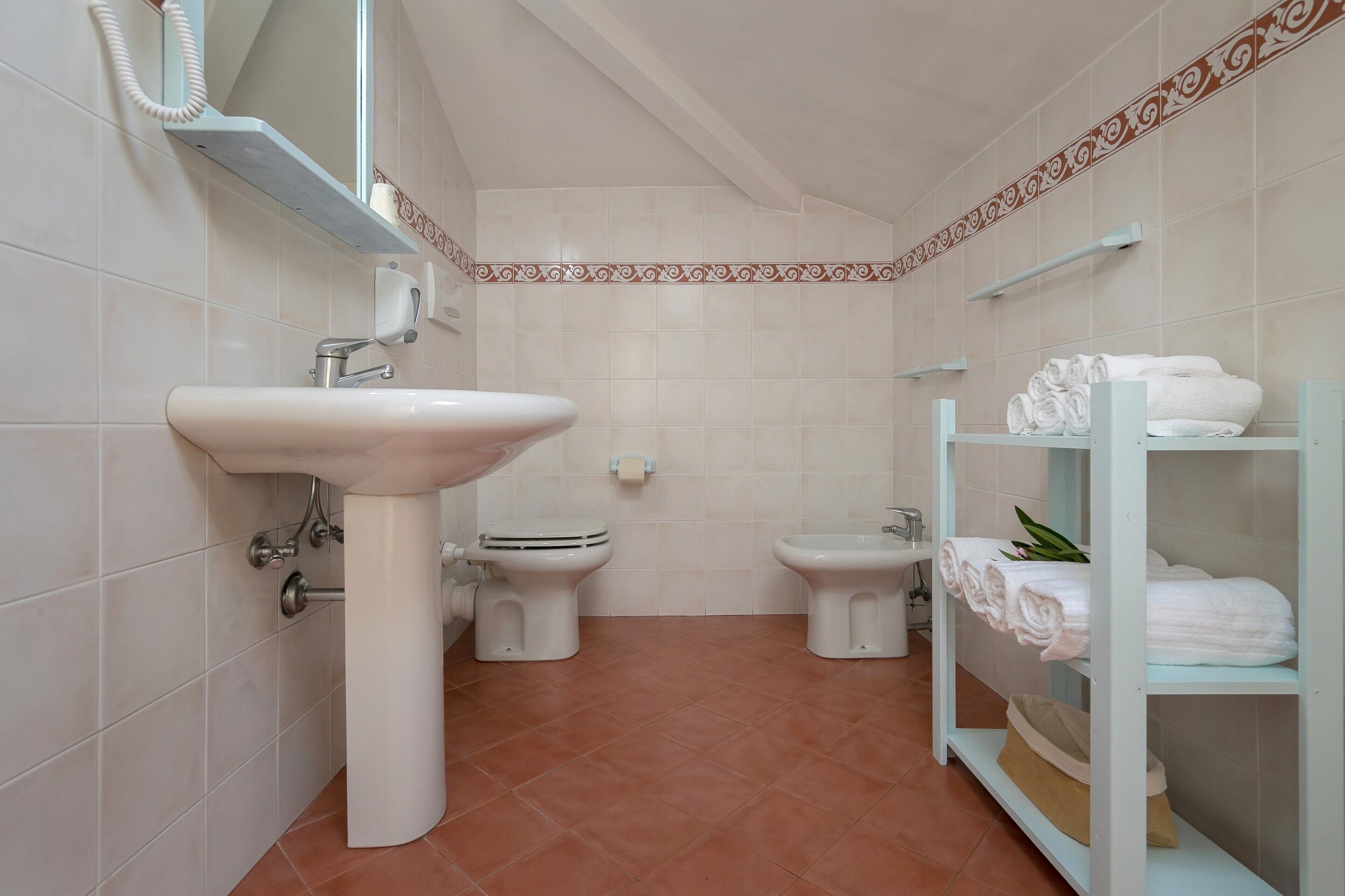 A bathroom with a sink, two toilets, a towel rack with towels, and a shelf with toiletries.