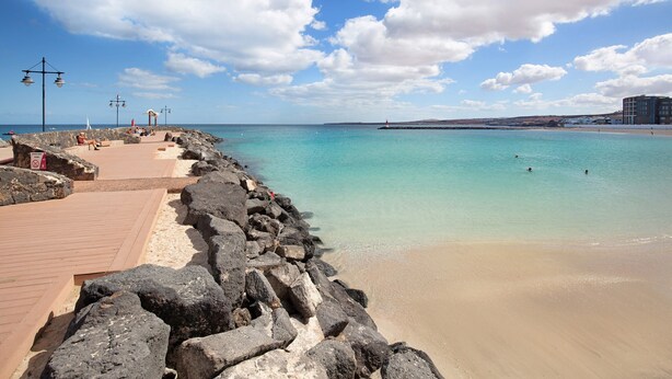 Un paseo marítimo con una barrera rocosa, una playa de arena y un cielo azul despejado.