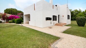 A white building with a paved entrance, a small porch, and a garden with pink flowers. - Holiday Home 'Pedras Del Rei Ll' with Shared Pool, Wi-Fi and Air Conditioning (Tavira)