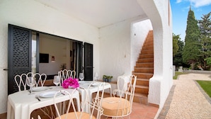 A dining area with a table set for four, white chairs, and a staircase leading to an upper level. - Holiday Home 'Pedras Del Rei Ll' with Shared Pool, Wi-Fi and Air Conditioning (Tavira)