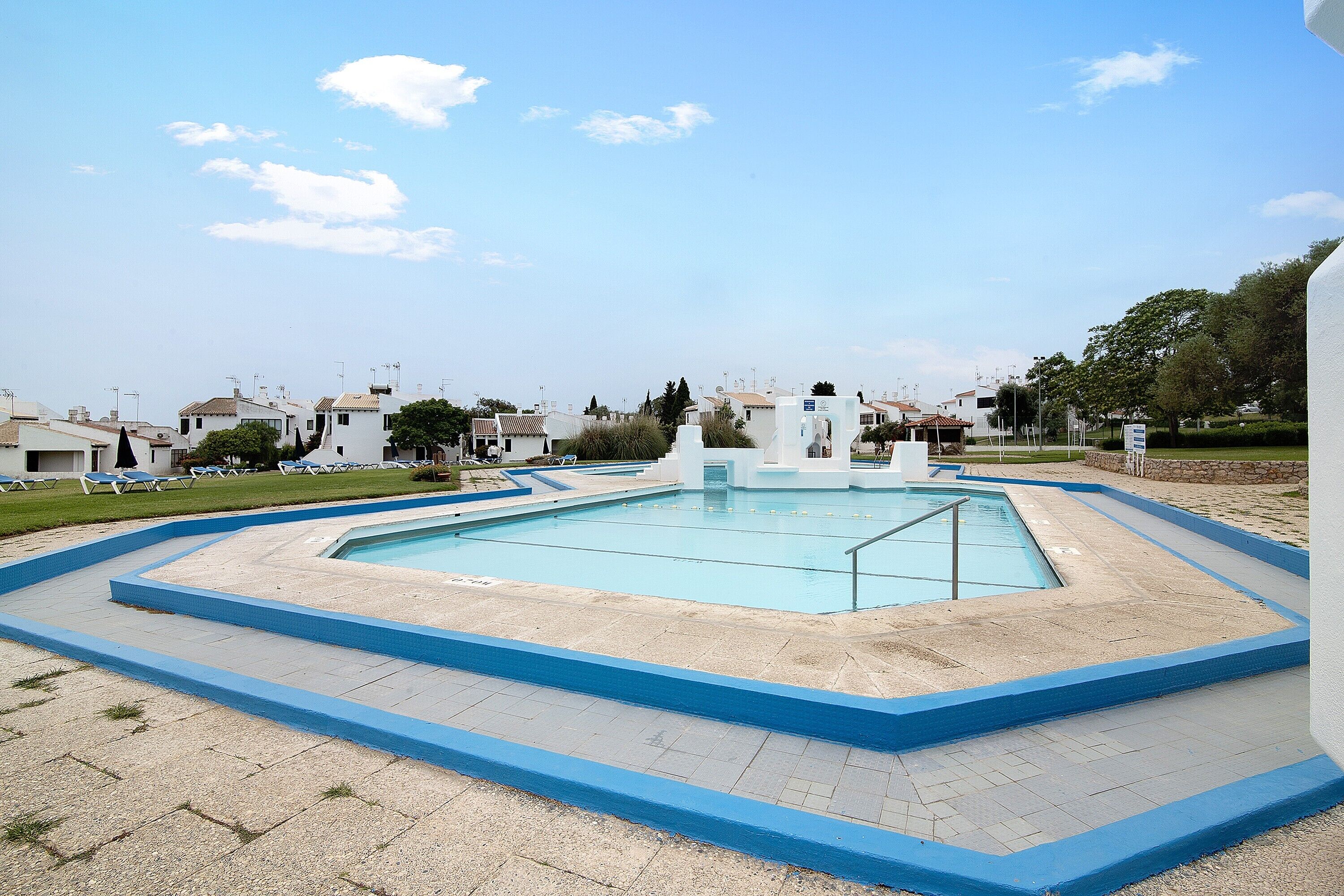 A swimming pool with a blue border, surrounded by a paved area with a few lounge chairs.