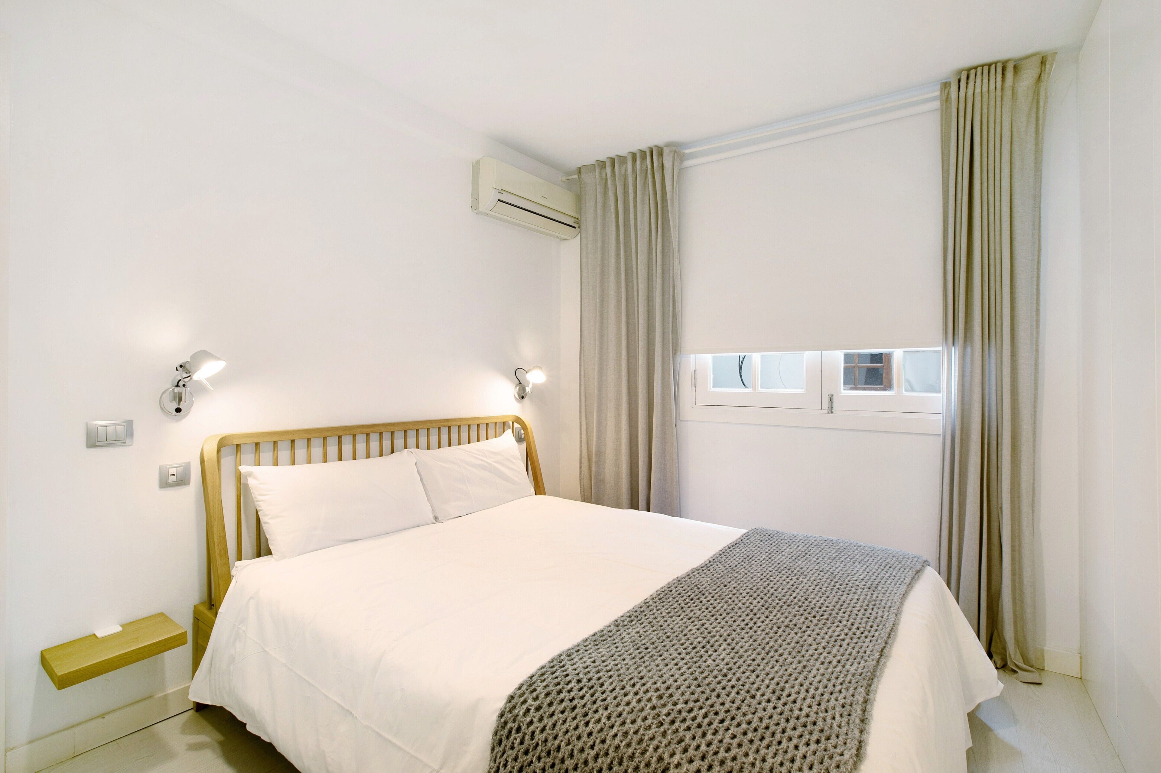 Une chambre à coucher comprenant un lit, des lampes de chevet, un appareil de climatisation, une fenêtre avec des rideaux et une petite étagère.