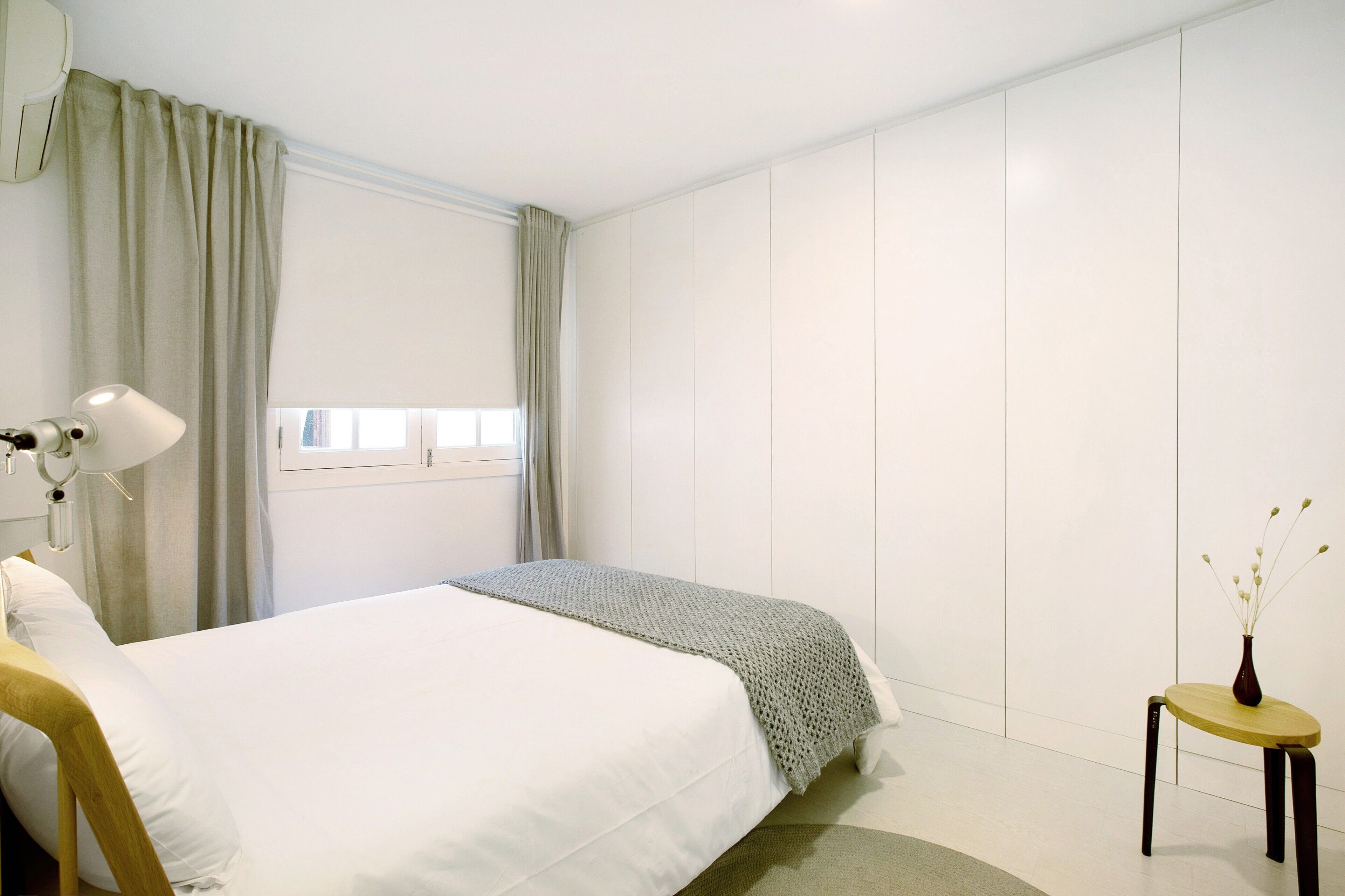 A bedroom with a bed, a bedside lamp, a small table with a vase, and a window with curtains.