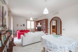 Ein Wohnzimmer mit einem weißen Sofa, einem Esstisch mit Stühlen, einem Bücherregal und einem Fernseher.