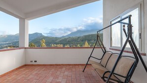 Ein Balkon mit einer Schaukel, gefliestem Boden und Blick auf grüne Hügel und Berge.