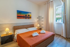 Une chambre à coucher comprenant un lit, deux tables de chevet, une commode, un élément de décoration murale en forme de croix, un tableau et une fenêtre avec des rideaux.