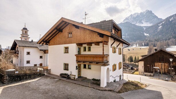 Un tipico chalet con balcone in legno, parete in pietra e montagne sullo sfondo.