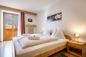 A bedroom with a wooden bed, white bedding, a nightstand with a lamp, a radiator, and a window with curtains.