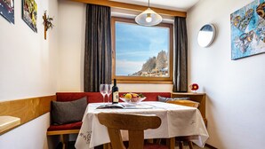 Un comedor con una mesa dispuesta para dos personas, un banco rojo, una silla de madera, un ventanal con vistas a árboles y edificios, y un cuadro en la pared.
