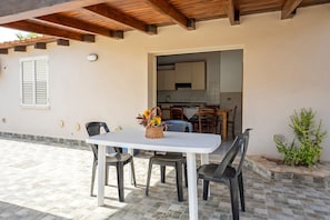 Une terrasse extérieure avec une table blanche et quatre chaises, un plafond en bois et une vue sur un espace cuisine.