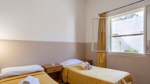 Ein Hotelzimmer mit zwei Betten, einem Nachttisch, einem Fenster mit Vorhängen und Blick auf Grünfläche.