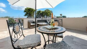 Une terrasse avec une table ronde, deux chaises, un grand parasol et une bouteille de champagne.