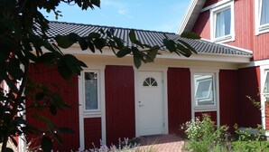 Ein rotes Gebäude mit weißer Tür und Fenstern, ein Ziegelweg, der zum Eingang führt, und umliegendes Grün.