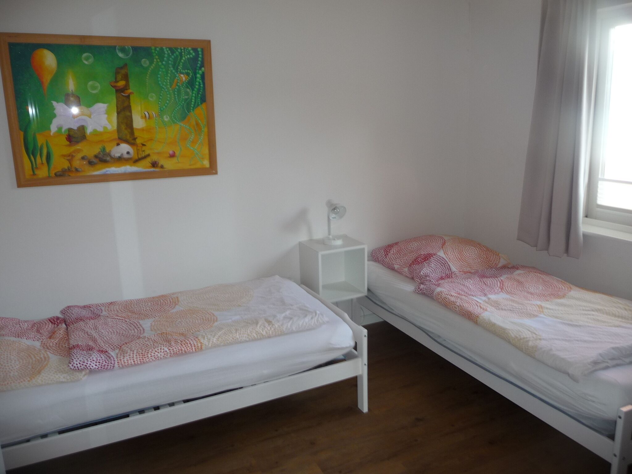 A room with two single beds, a nightstand with a lamp, a painting on the wall, and a window with curtains.