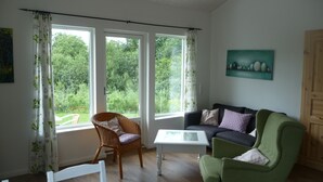 Ein Wohnzimmer mit einem grünen Sessel, einer dunkelgrauen Couch, einem Korbsessel und einem weißen Couchtisch. Es gibt drei Fenster mit Vorhängen, ein Bild an der Wand und eine Holztür.