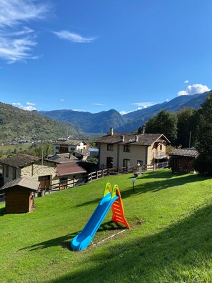 A playground with a slide and climbing structure in a grassy area with houses and mountains in the background. - Apartment 'Il Giardino' with Mountain View, Private Terrace and Wi-Fi (Castello dell'Acqua)