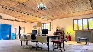 Ein Zimmer mit einem reetgedeckten Dach, einem Schreibtisch mit Computer, einem Stuhl, einem Tisch, einer blauen Tür und einem Fenster mit Blick auf Bäume.