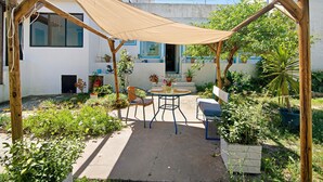 Eine hölzerne Pergola mit beigem Sonnensegel, einem runden Tisch, zwei Stühlen und Kübelpflanzen in einem Innenhof.