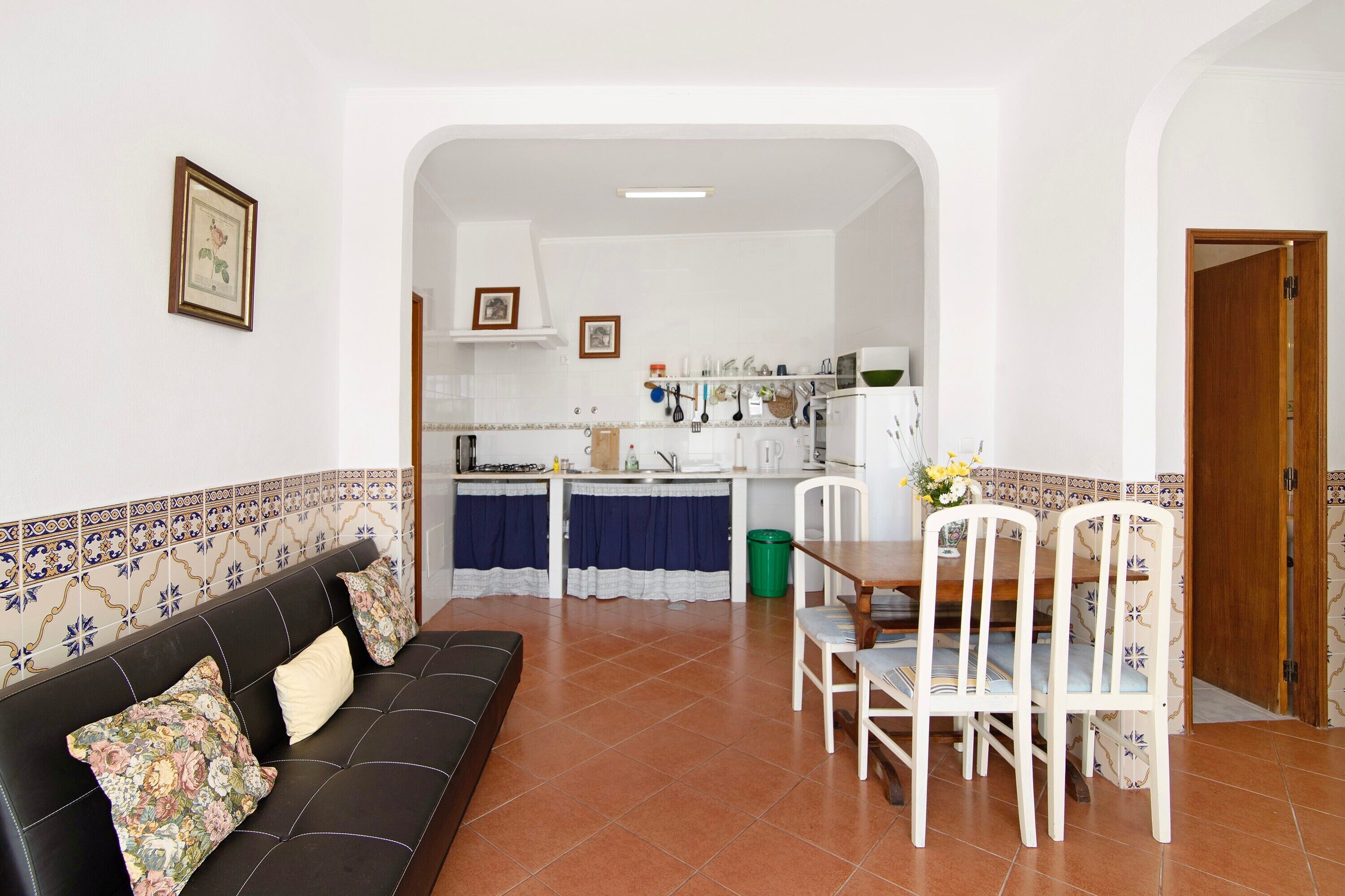 A living room with a sofa, dining table, chairs, and a kitchen in the background.