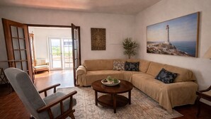 Ein Wohnzimmer mit einer Couch, einem Sessel, einem Couchtisch, einem Gemälde eines Leuchtturms und einer Tür, die zu einem Außenbereich führt.