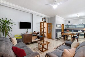 Uma sala de estar com sofá, mesa de centro, televisão, área de jantar com cadeiras e cozinha ao fundo.
