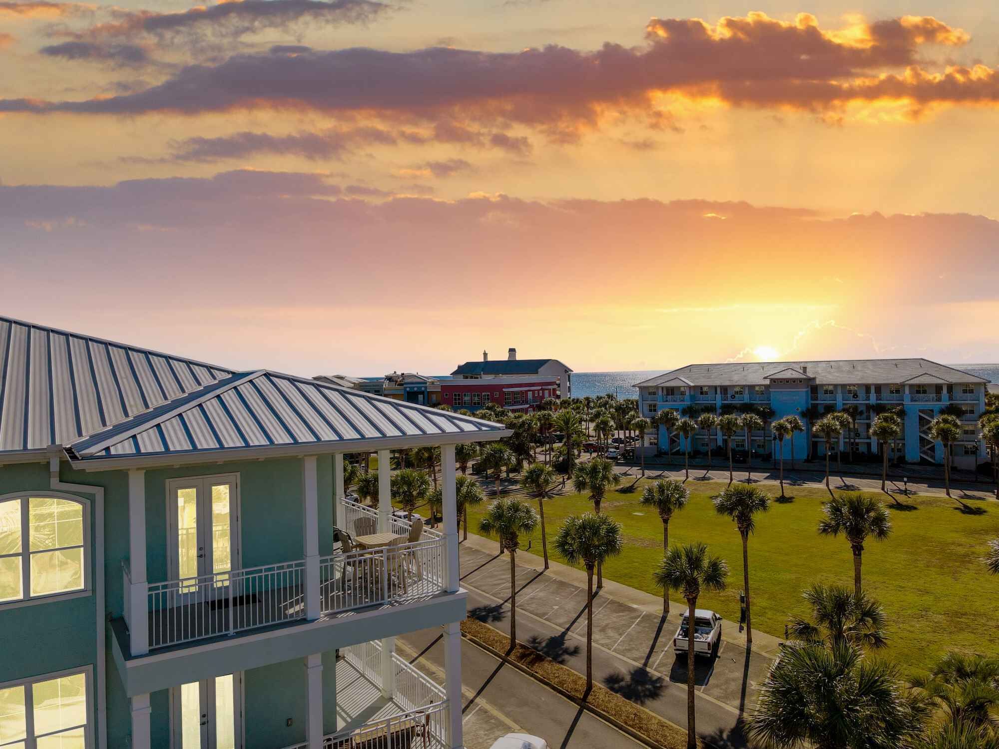 A multi-story building with a balcony, surrounded by palm trees and a parking lot, overlooking a scenic sunset.