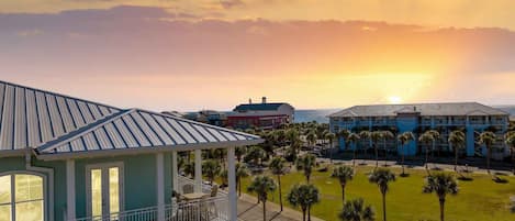 A multi-story building with a balcony, surrounded by palm trees and a parking lot, overlooking a scenic sunset.