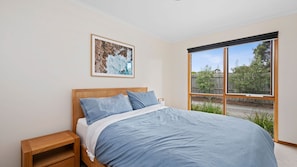 A bedroom with a bed, a nightstand, a window with a view of greenery, and a framed picture on the wall. - Roadknight Ocean Road View 236 (Anglesea)