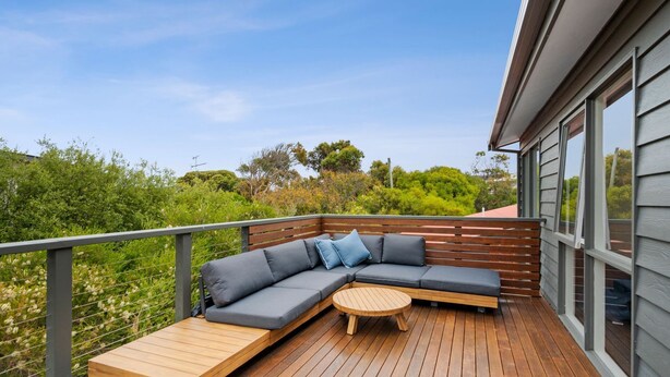 A deck with a wooden floor, a grey sectional sofa with blue cushions, and a round wooden table. - Roadknight Ocean Road View 236 400m to Beach (Anglesea)