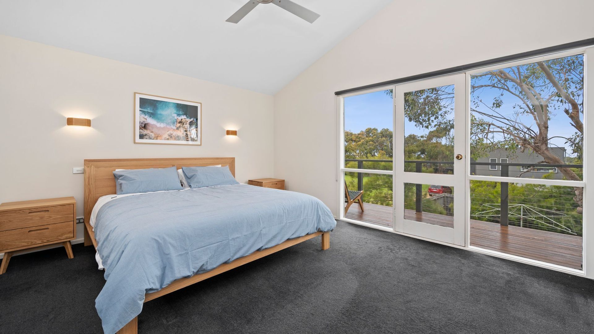 A bedroom with a bed, bedside tables, a ceiling fan, a large window with a view of greenery, and a framed picture on the wall.