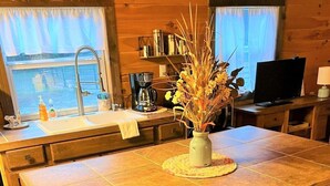 Interior de una cabaña de madera con una zona de cocina que incluye un lavamanos, cafetera y un florero con flores secas sobre una mesa.