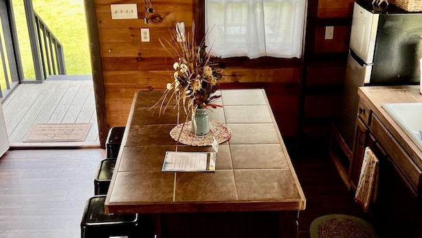 Un interior de madera con una mesa de comedor, sillas, un florero con flores, un refrigerador y una ventana con cortinas.