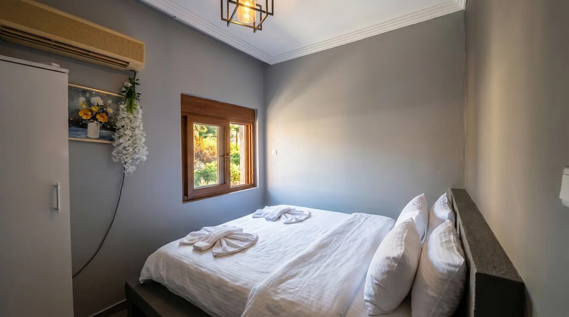 A bedroom with a bed, a window, a wall-mounted air conditioner, and a hanging floral arrangement.