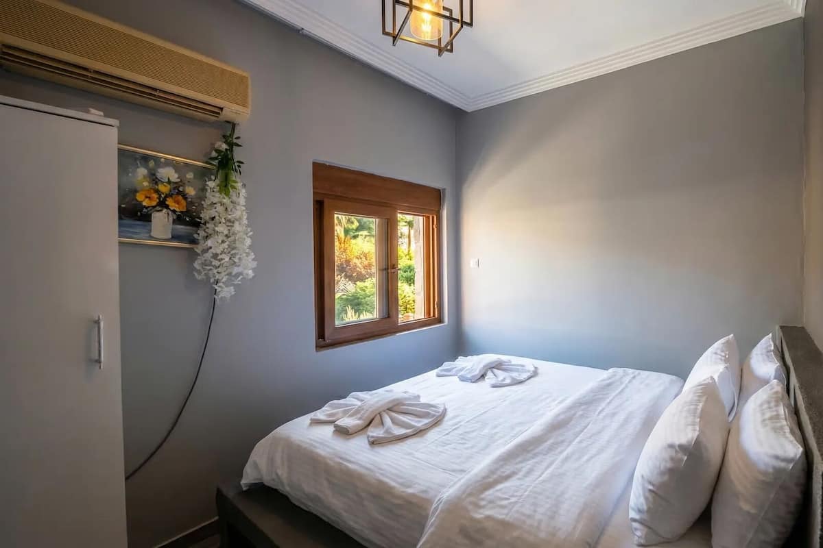 A bedroom with a bed, a window, a wall-mounted air conditioner, and a hanging floral arrangement.