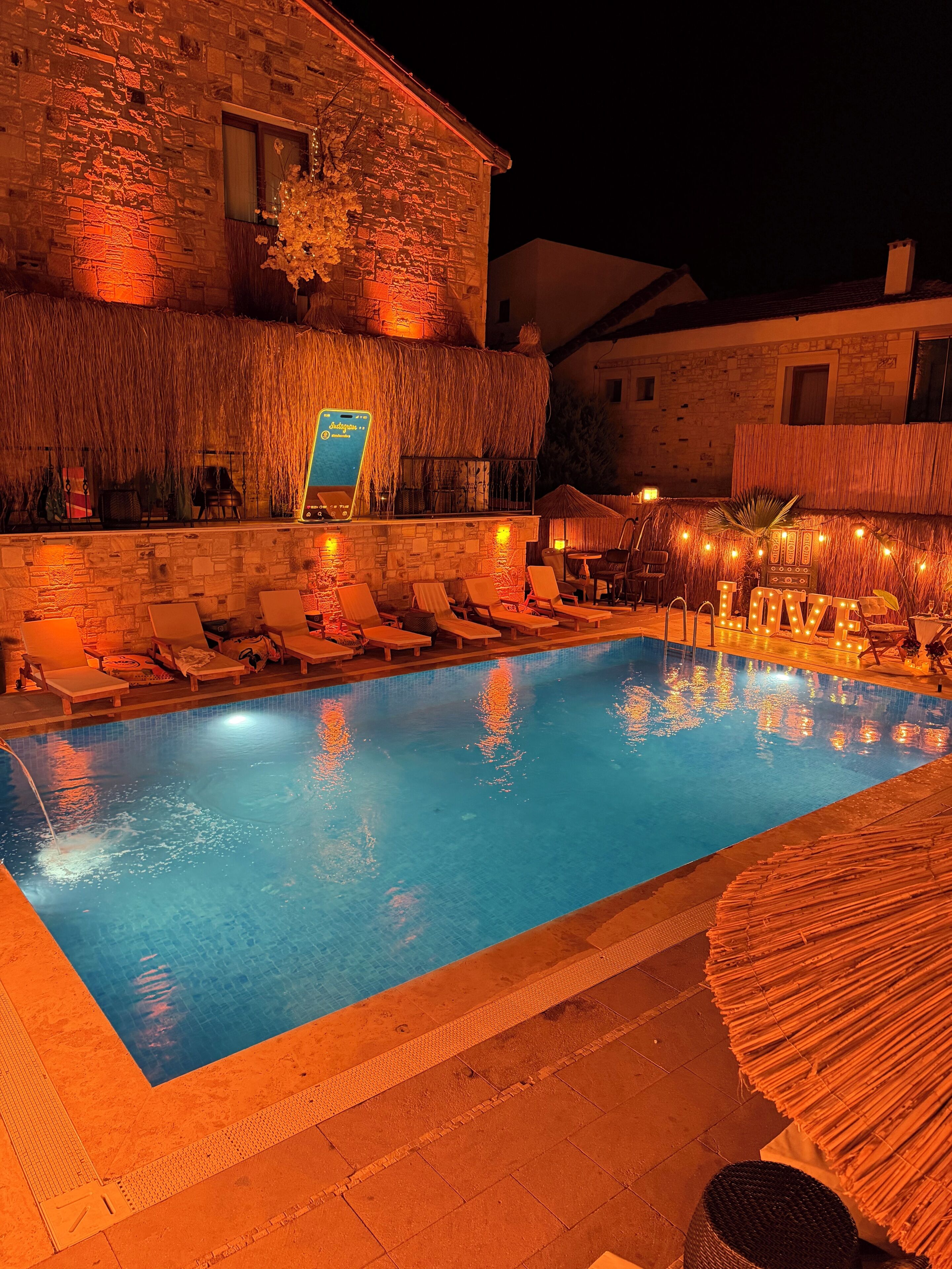 A swimming pool with lounge chairs, a thatched roof structure, and a stone building in the background.