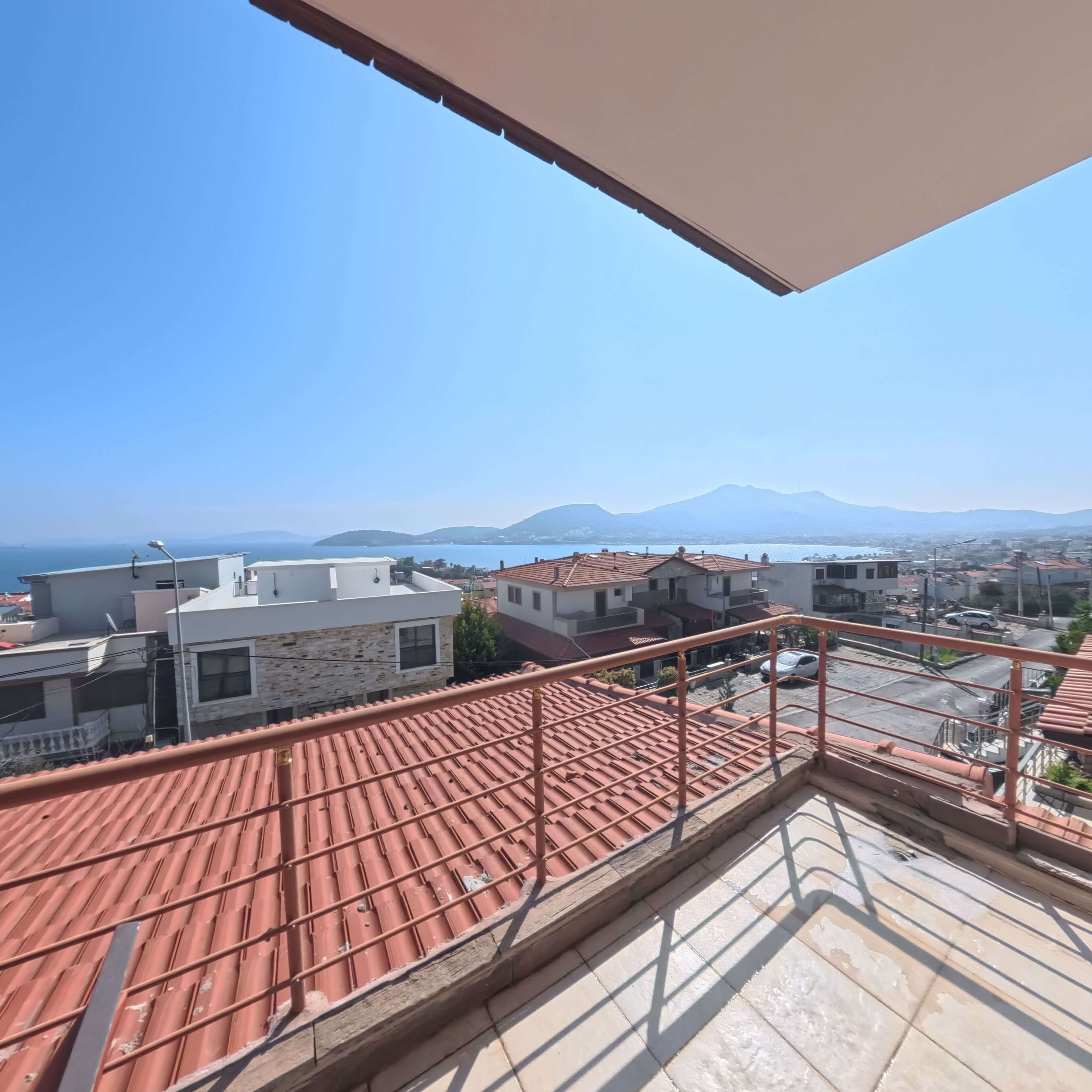 A balcony with a red-tiled roof overlooking a residential area with houses and a distant mountain range.