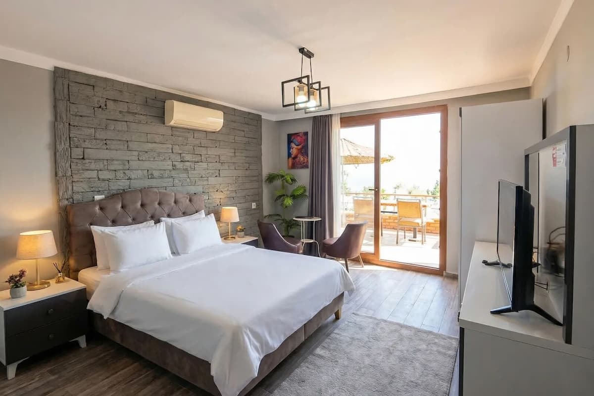 A modern hotel room with a large bed, a TV, a desk, and a balcony with outdoor seating.