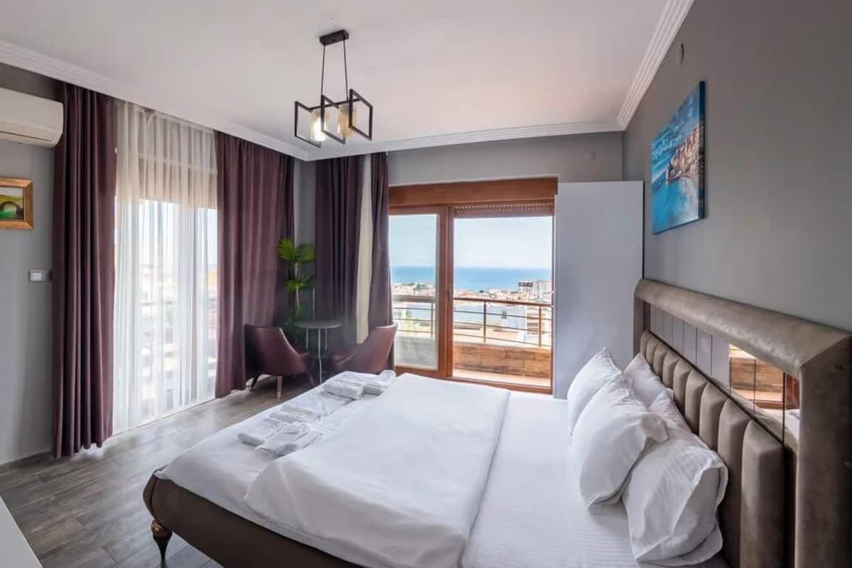 A hotel room with a bed, a desk, a chair, a plant, and a view of the city.