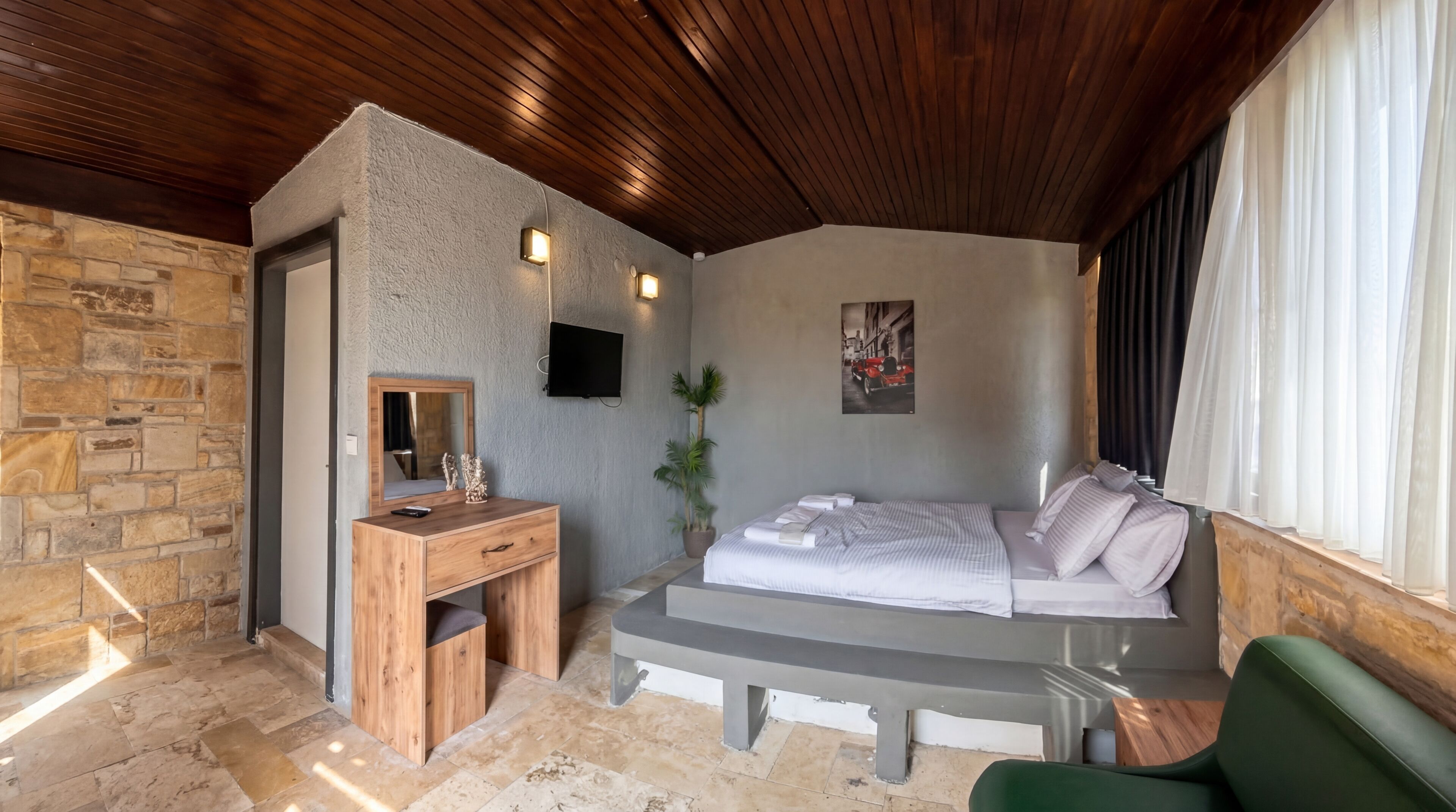 Quarto com parede de pedra, teto de madeira, uma cama, escrivaninha, espelho, planta em vaso e televisão.
